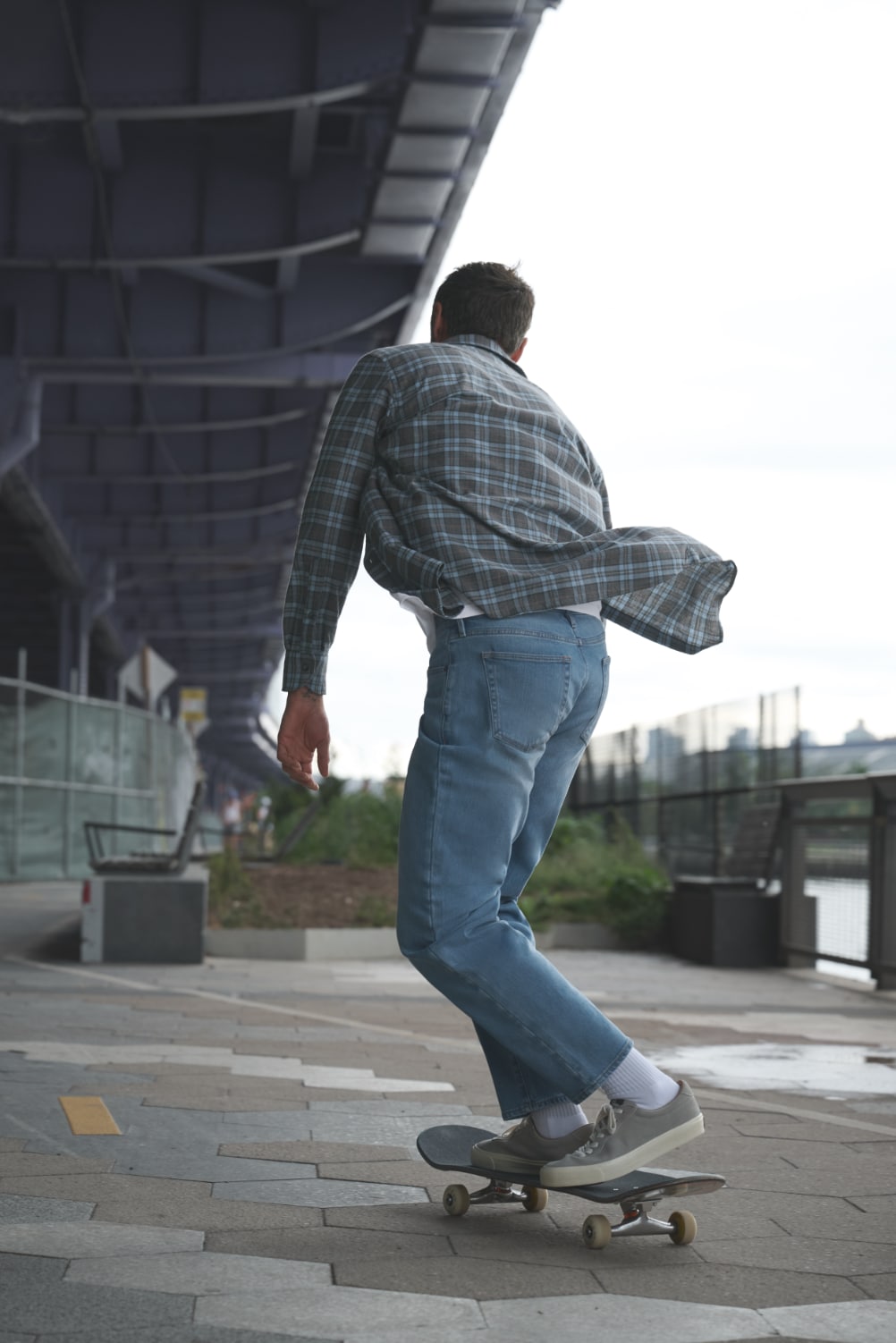 Man skating away from camera, under highway bridge, shirt blowing in the wind