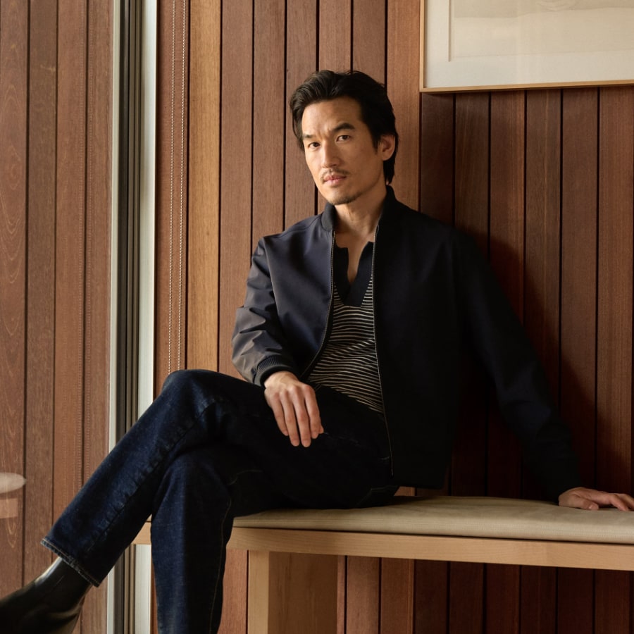 Man wearing knitwear with jacket and jeans against a wood paneled wall