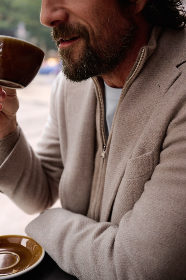 Man wearing a fall sport coat layered over a sweater, drinking coffee.